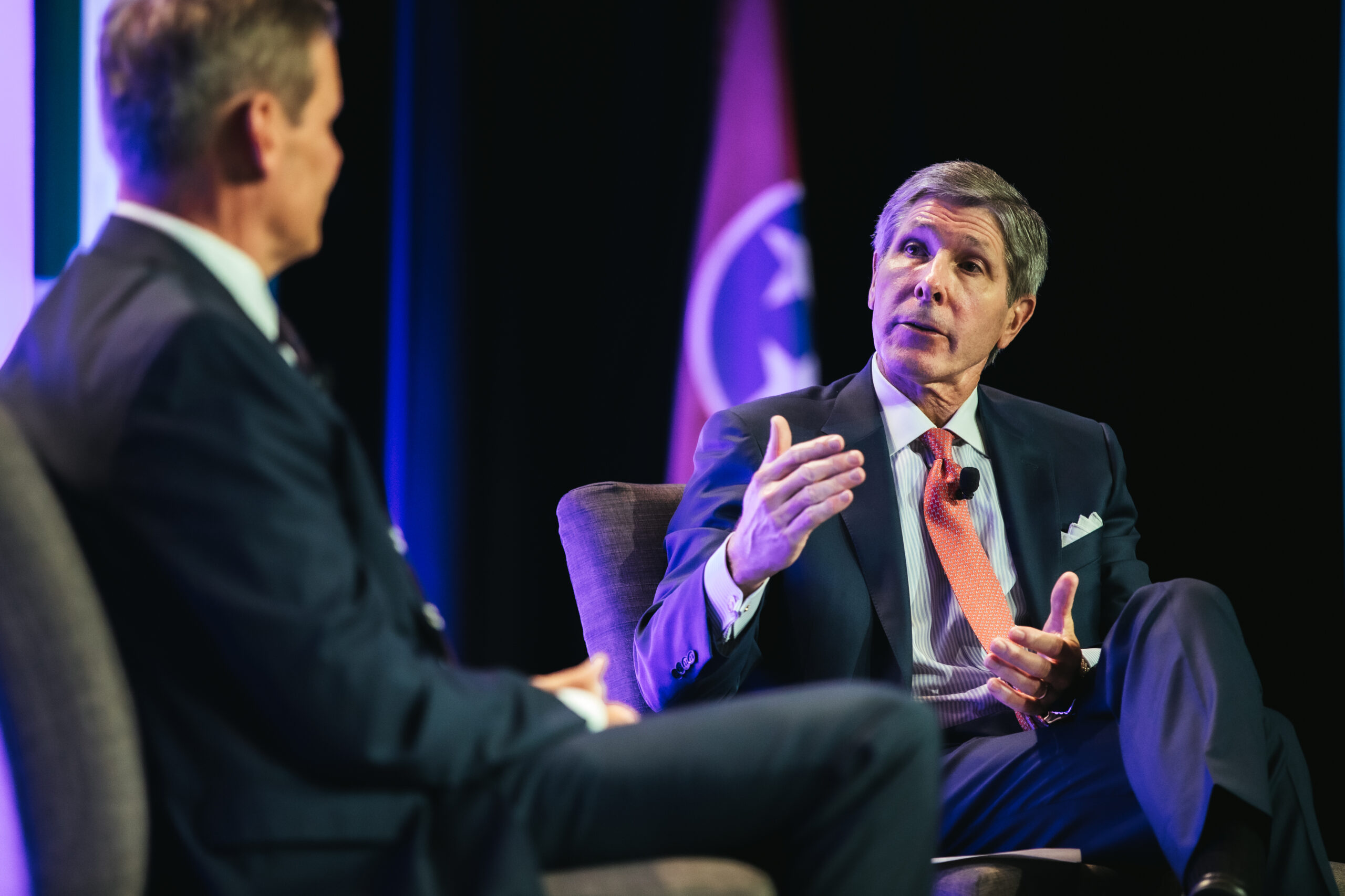 Two men in suits sit and talk on stage, one gesturing with his hand while speaking. A flag with a circular emblem is visible in the background. The setting appears formal, likely a conference or panel discussion.