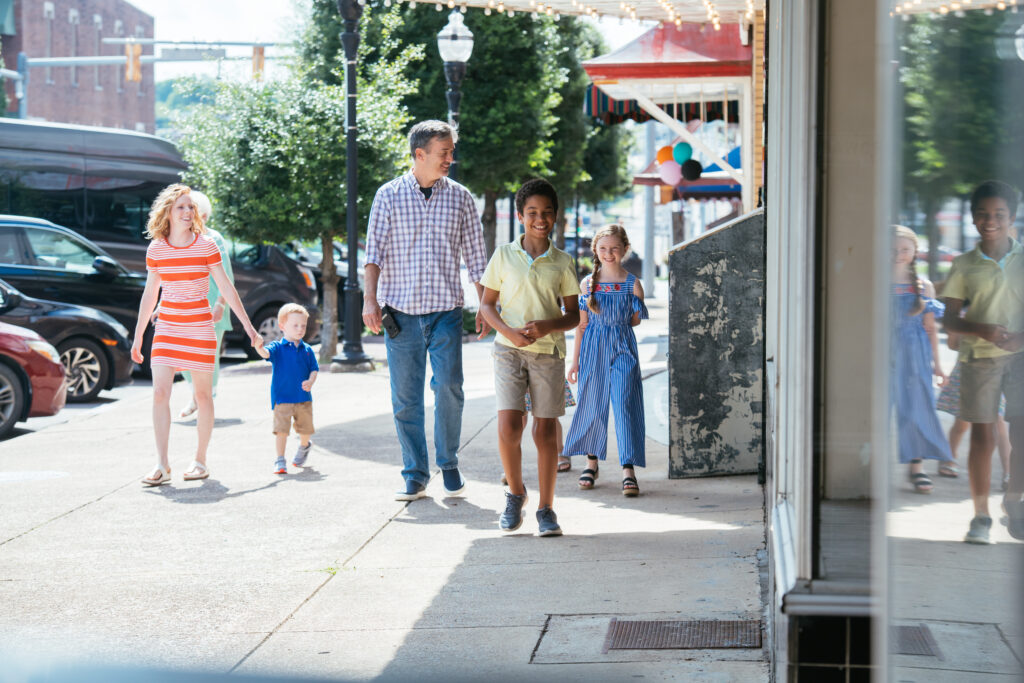A family with three children walks together on a sunny city sidewalk. The parents and kids smile as they approach a storefront, with trees and street signs in the background.