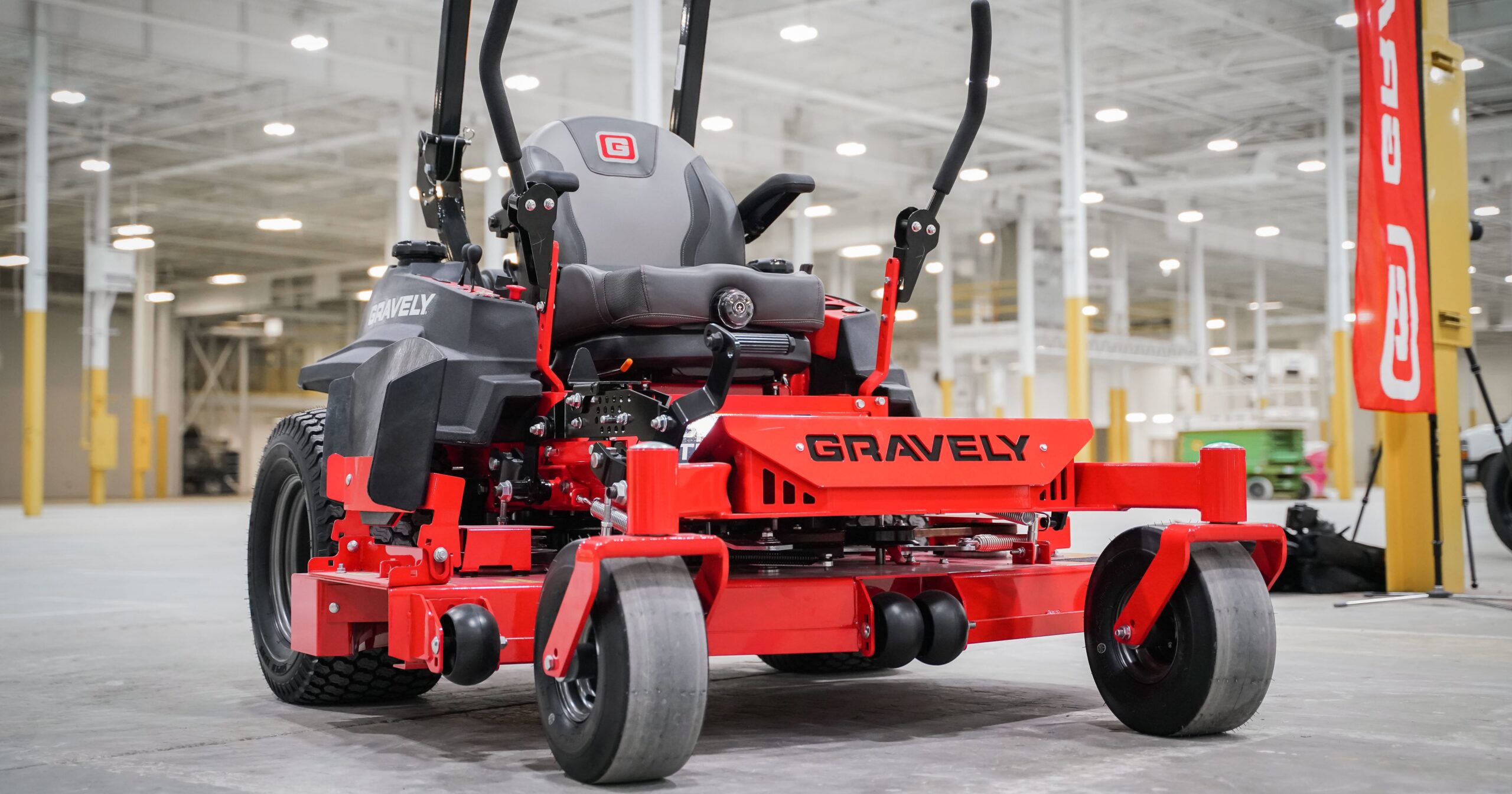 A red Gravely zero-turn lawn mower is parked indoors on a concrete floor in a large, well-lit industrial facility. The mower features a black seat, control levers, and thick, treaded rear tires.