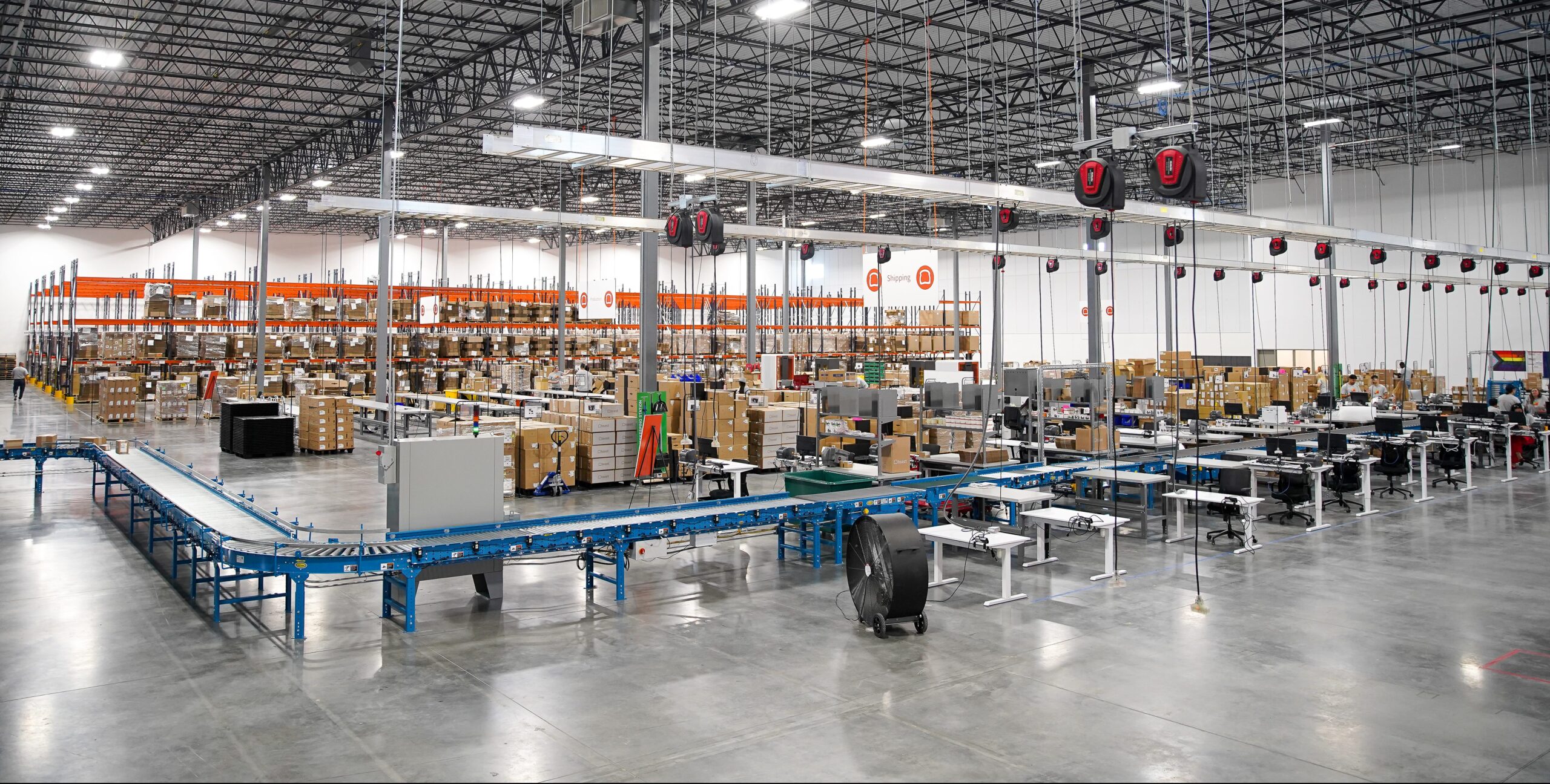 A spacious warehouse with high ceilings, shelves stacked with boxes, and a blue conveyor belt running through the center. Workstations and equipment are neatly arranged on the polished concrete floor.