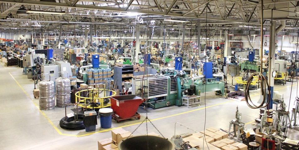 A large industrial factory floor filled with various machines, equipment, and workstations, with boxes, metal containers, and tools organized throughout the brightly lit space. No workers are visibly present.