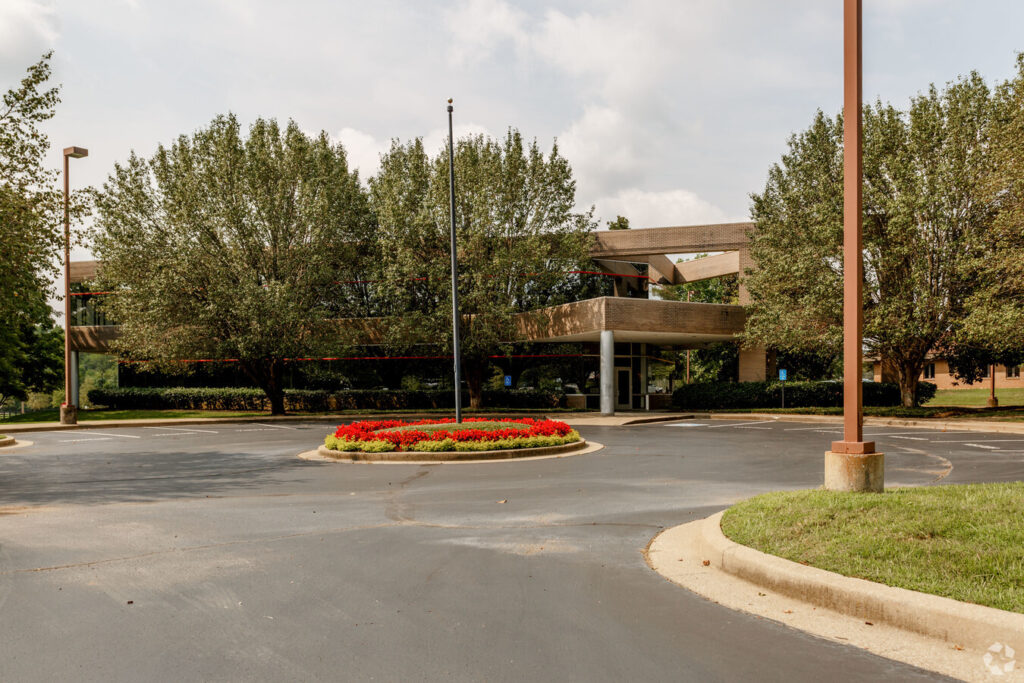 An empty circular driveway with a central flower bed and flagpole leads to a two-story office building partially obscured by trees on a cloudy day.