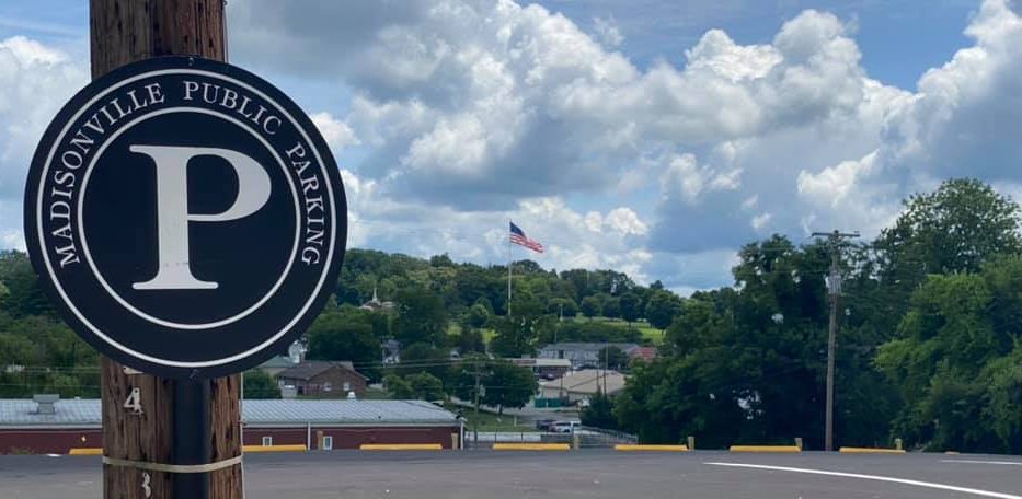A round sign on a wooden post reads "Madisonville Public Parking." In the background, there are trees, buildings, and a large American flag, all under a partly cloudy sky.