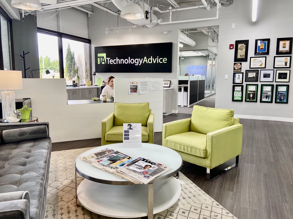 Modern office lobby with green armchairs, a round coffee table with magazines, gray sofa, and a reception desk. The wall behind the desk has a TechnologyAdvice sign and a gallery wall displays framed awards and photos.