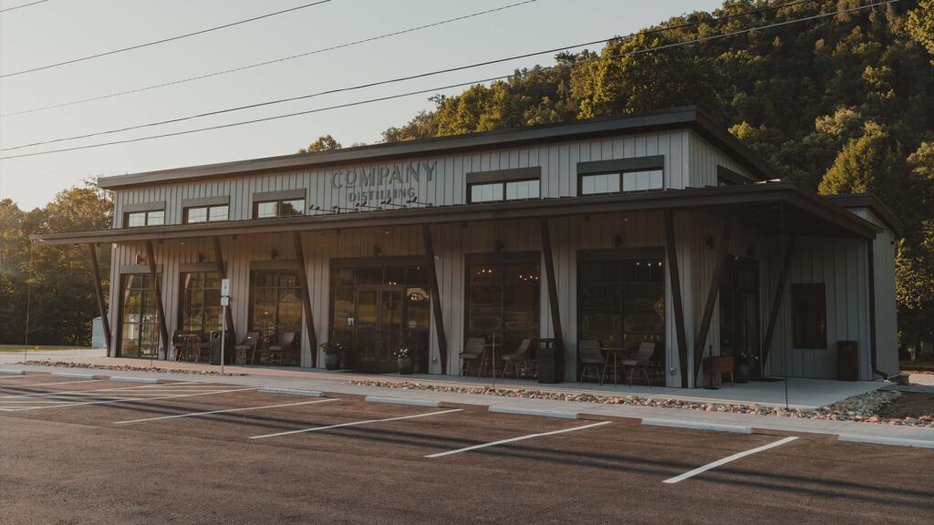 A modern, gray metal building with large windows and an overhanging roof, labeled “Company Distilling,” sits beside an empty parking lot and lush, green trees on a sunny day.