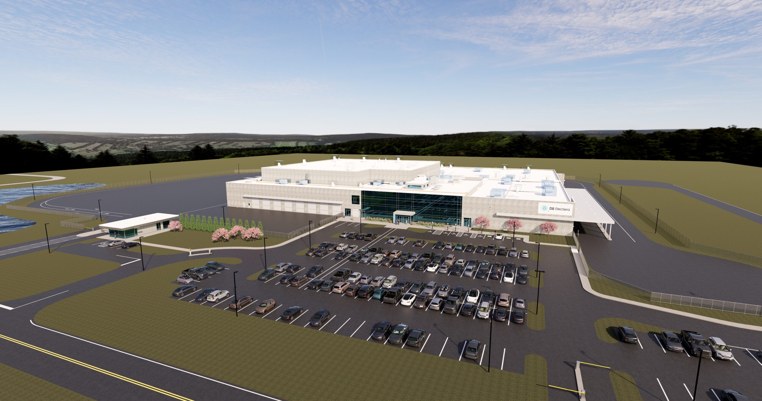 Aerial view of a large, modern industrial building with a glass entrance, surrounded by parking lots filled with cars, fences, green lawns, and trees in the background under a partly cloudy sky.