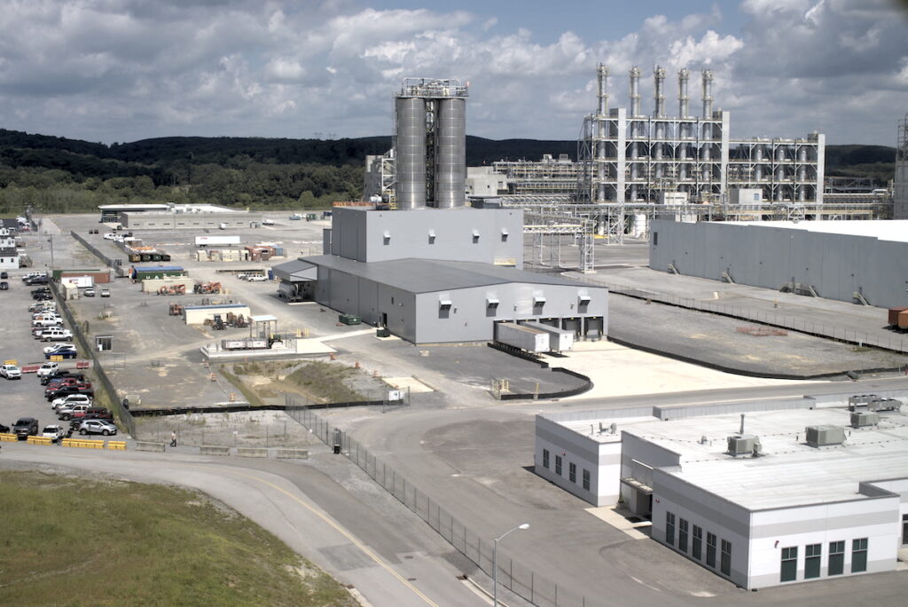 An industrial complex with multiple large factory buildings, tall silos, and metal structures surrounded by paved lots, parked vehicles, and security fencing, set against a backdrop of trees and cloudy sky.