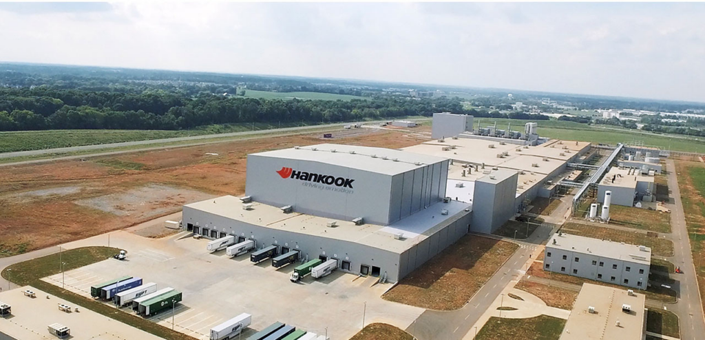 Aerial view of a large Hankook manufacturing facility with multiple loading docks and trucks parked outside, surrounded by open land and trees in the background.