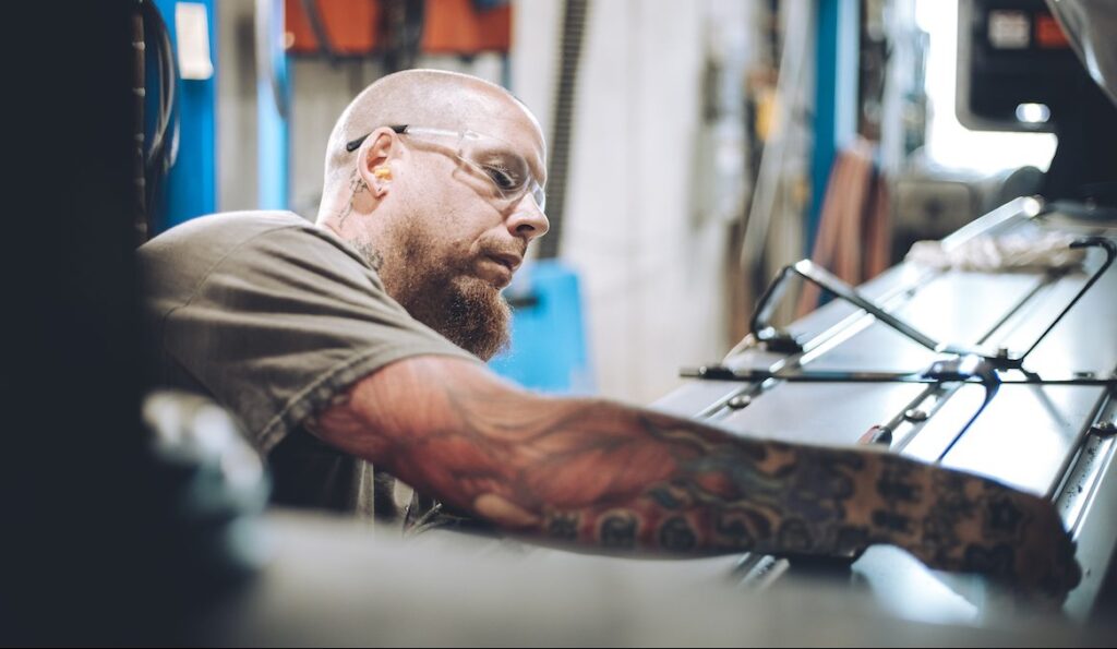 A man with a beard, safety glasses, and colorful tattoos works intently on a piece of industrial machinery in a workshop setting.