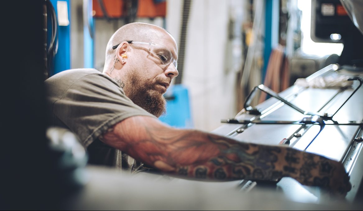 A man with a beard, safety glasses, and colorful tattoos works intently on a piece of industrial machinery in a workshop setting.