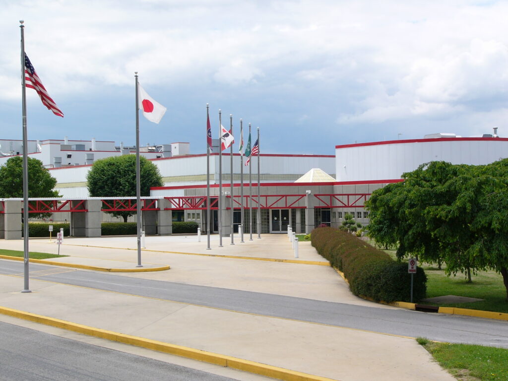 A modern building with a curved facade, multiple flagpoles displaying various international flags, a driveway with yellow curbs, and trimmed bushes and trees under a partly cloudy sky.