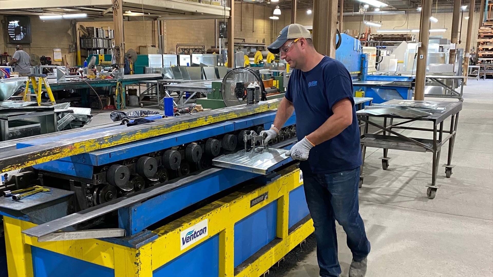 A man wearing a cap, glasses, gloves, and blue shirt operates a large metal bending machine in a spacious industrial workshop filled with equipment and metal sheets.