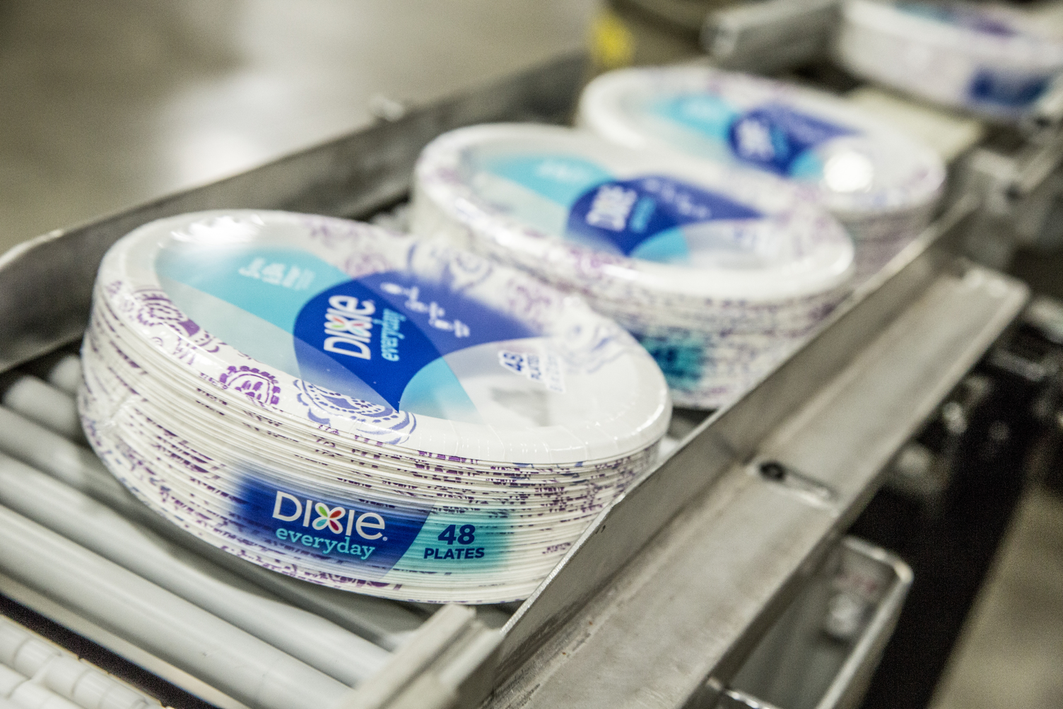 Stacks of packaged Dixie Everyday paper plates, each pack containing 48 plates, move along a conveyor belt in what appears to be a manufacturing or packaging facility.