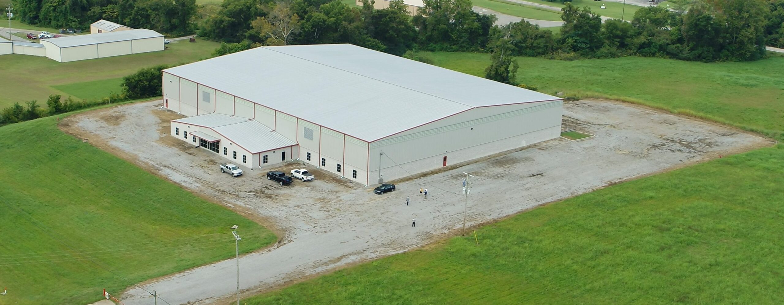 Aerial view of a large, white industrial warehouse with a metal roof, surrounded by a gravel lot and grassy fields, with a few vehicles and people near the building’s entrance.