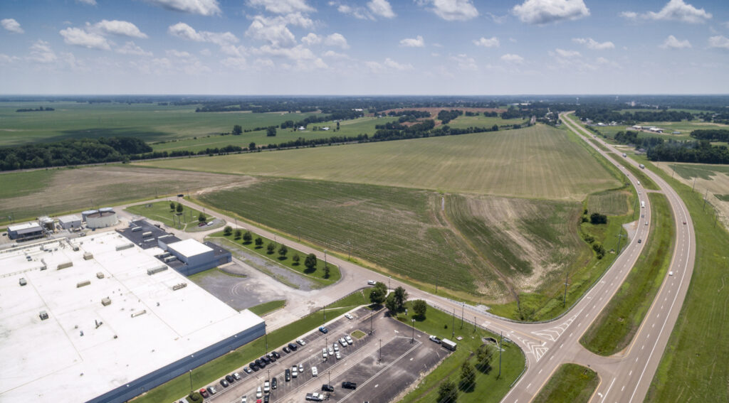 Aerial view of a large industrial building with a white roof, a parking lot with cars, surrounding green fields, and a highway curving through the rural landscape under a partly cloudy sky.