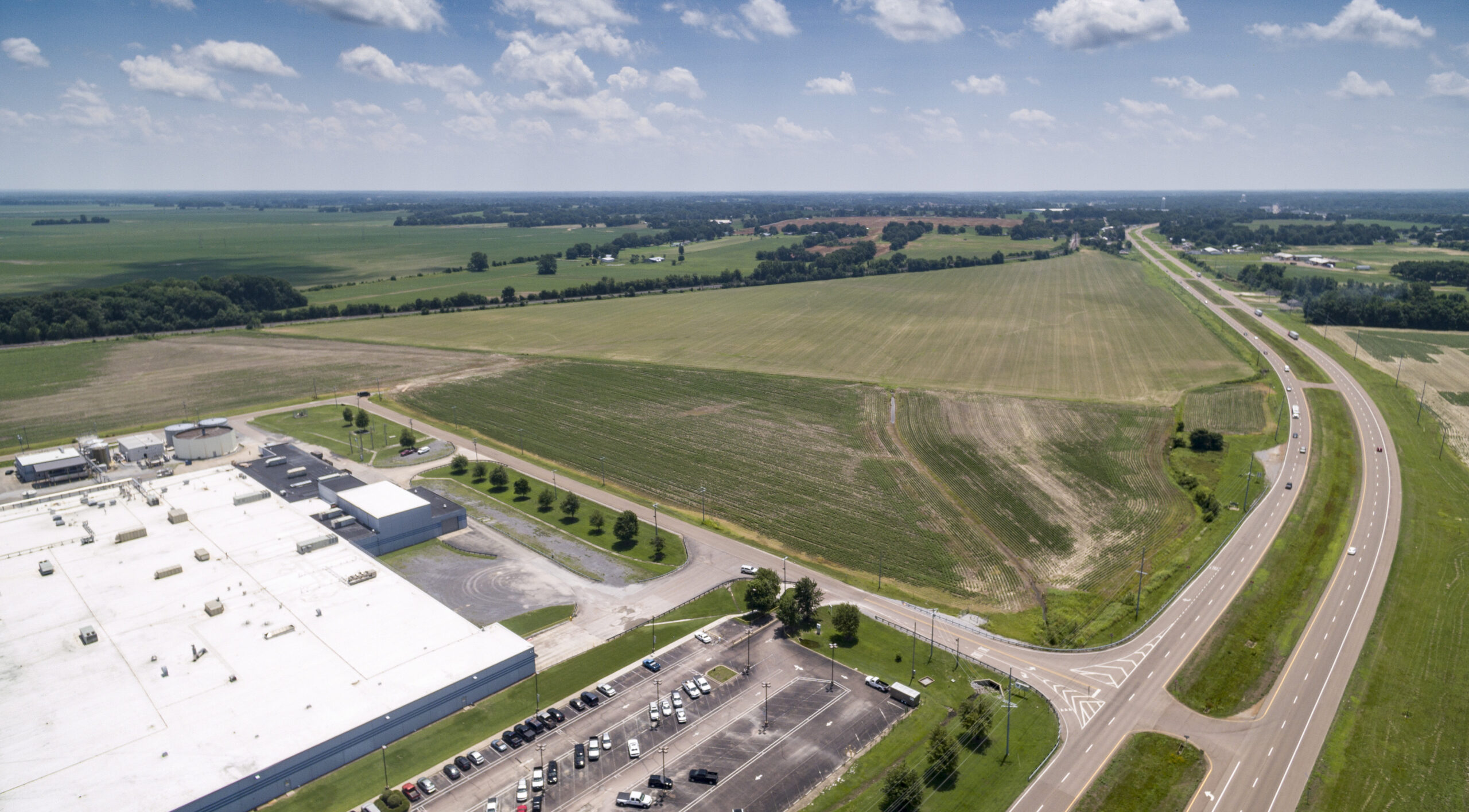 Aerial view of a large industrial building with a white roof, a parking lot with cars, surrounding green fields, and a highway curving through the rural landscape under a partly cloudy sky.