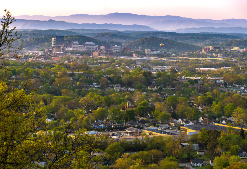 A scenic view of a city nestled among green trees, with several buildings in the distance and mountains layered in the hazy background under a pastel sky.
