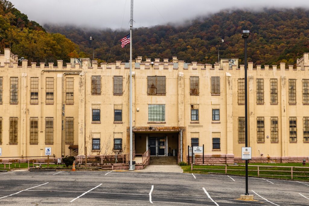 A large, beige prison building with barred windows stands in front of a parking lot. An American flag flies on a pole, and misty, tree-covered hills rise in the background.