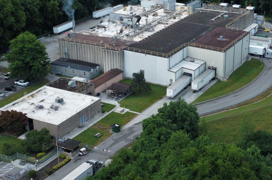 Aerial view of an industrial facility with two large buildings, several trucks parked at loading docks, and surrounding greenery, including trees and grass. Road curves around the buildings. Steam or smoke rises from the rooftop.