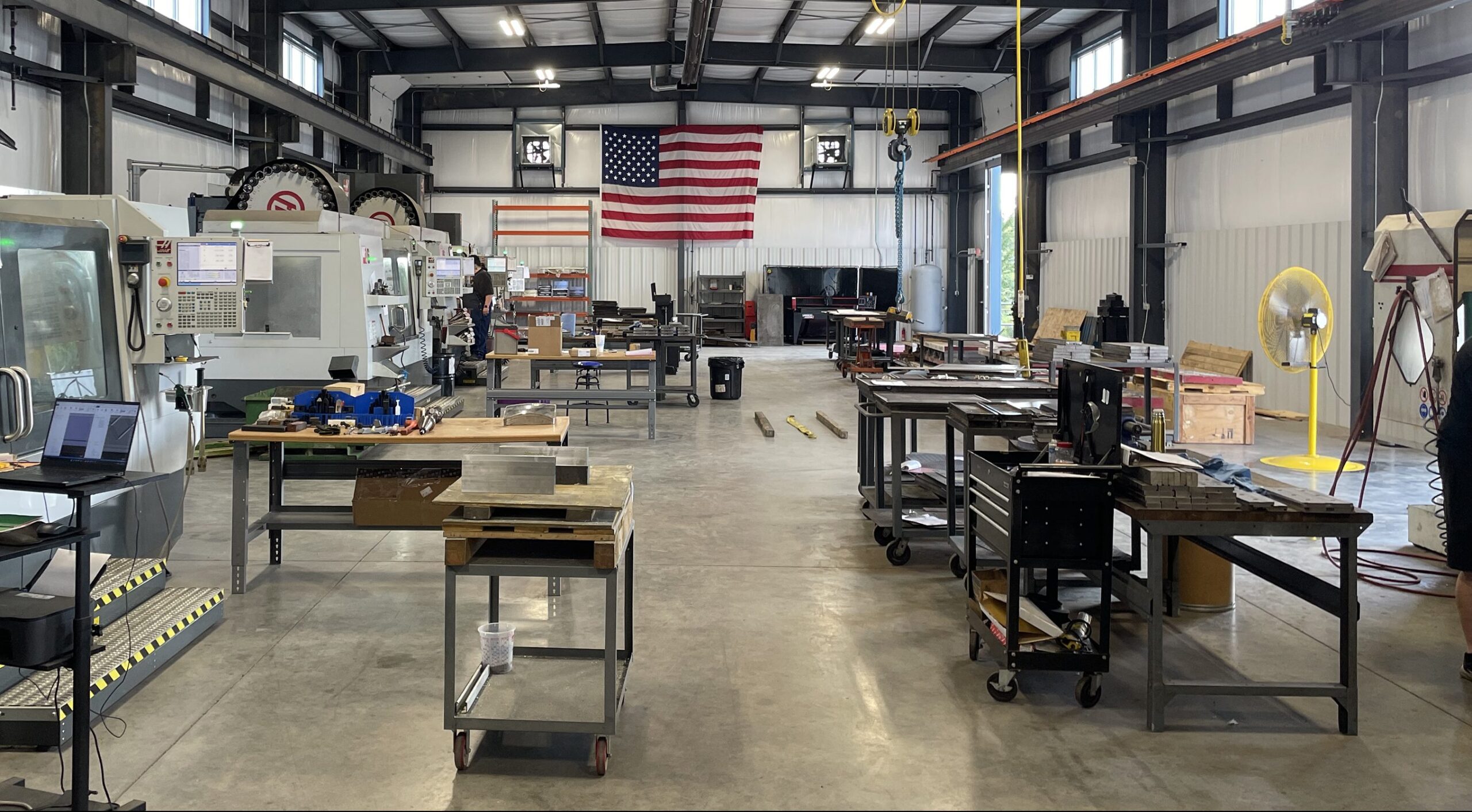 A spacious industrial workshop with various workstations, machinery, and tools. The floor is concrete, and an American flag hangs on the back wall. The area is clean, organized, and well-lit with natural and artificial light.