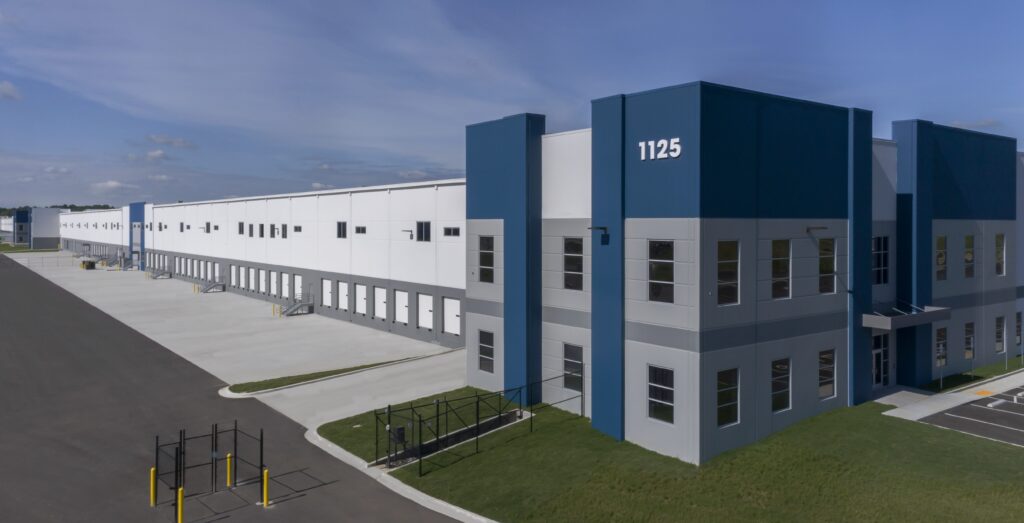 Large, modern industrial warehouse with blue and gray accents, marked “1125.” The building has multiple loading docks and windows, sits beside an empty paved lot, and is surrounded by green grass under a blue sky.