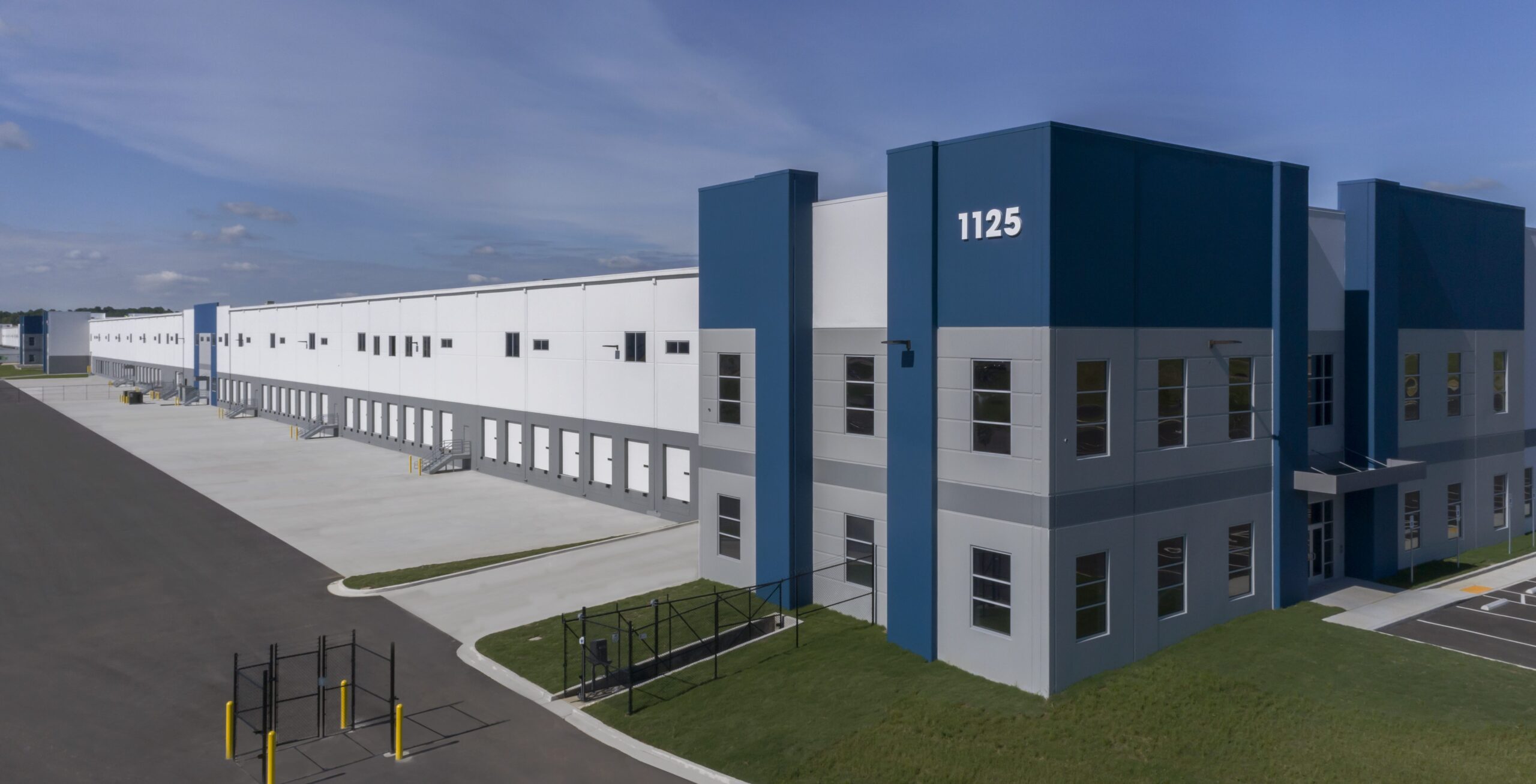 Large, modern industrial warehouse with blue and gray accents, marked “1125.” The building has multiple loading docks and windows, sits beside an empty paved lot, and is surrounded by green grass under a blue sky.
