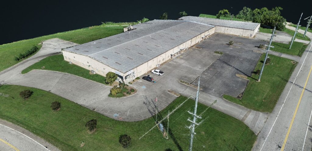 Aerial view of a large, rectangular industrial building with a gray roof, surrounded by a parking lot, grassy areas, a few parked cars, and utility poles along adjacent roads.