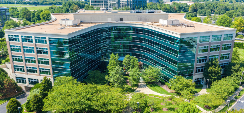 A modern, glass-fronted office building with a curved facade is surrounded by lush green trees and landscaped paths, viewed from above on a sunny day.
