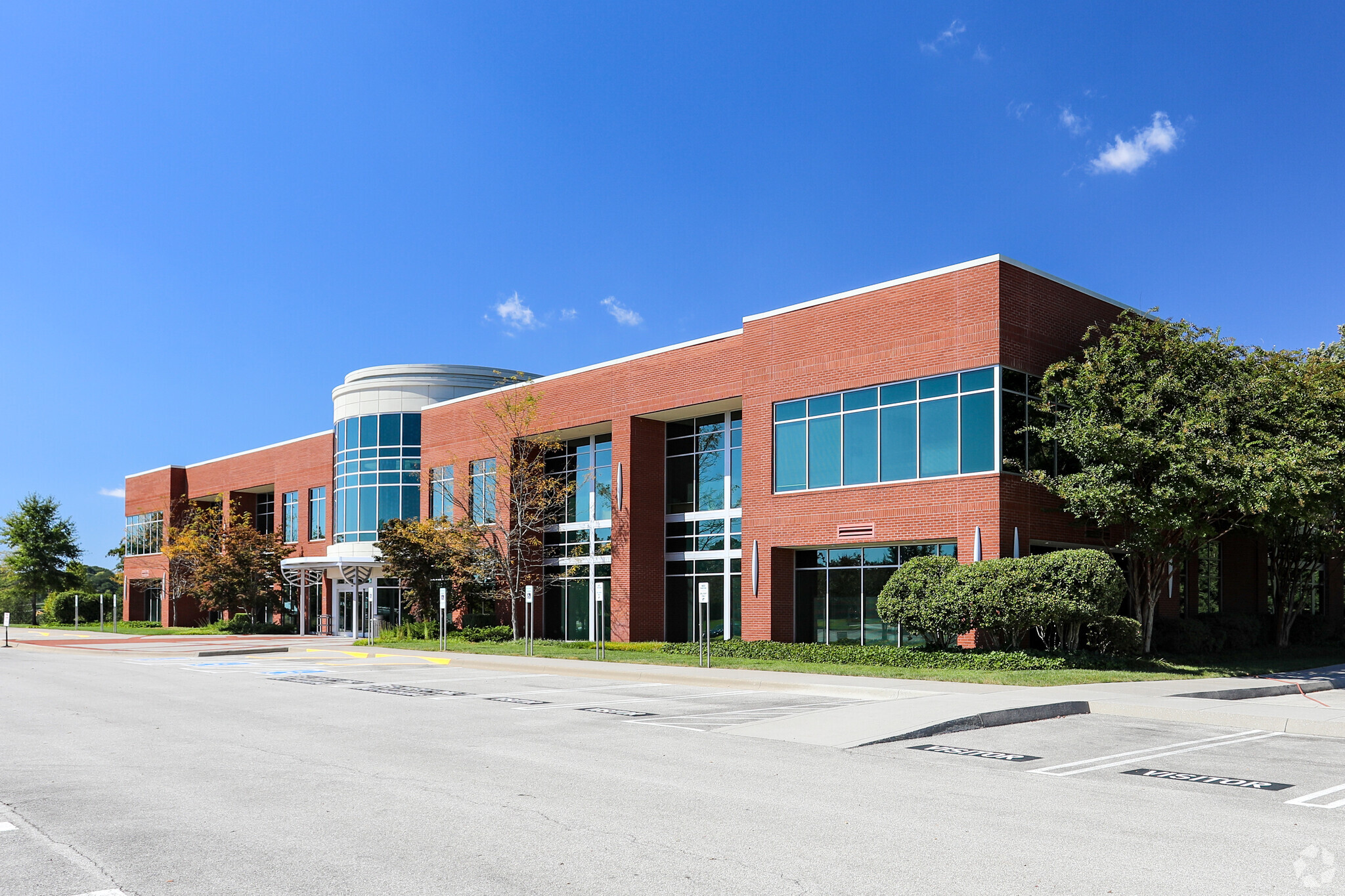 A modern two-story brick office building with large glass windows, surrounded by trees and greenery, under a clear blue sky. The building has a circular glass entrance and an empty parking lot in front.
