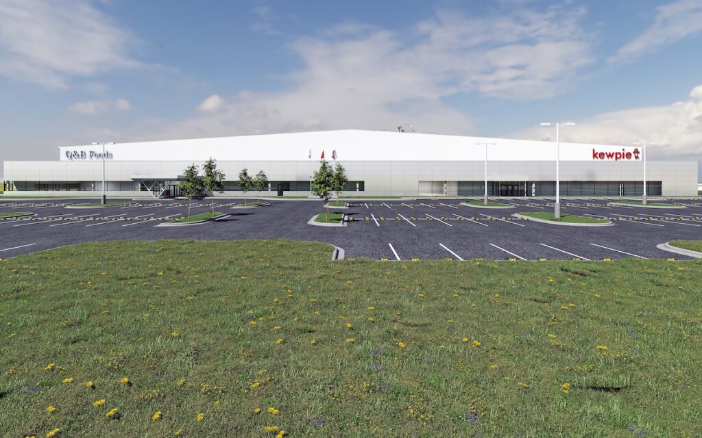 A large, modern industrial building with “Eagle Foods” and “kewpie” signs, a mostly empty parking lot, few trees, and a grassy field with yellow flowers in the foreground beneath a partly cloudy sky.