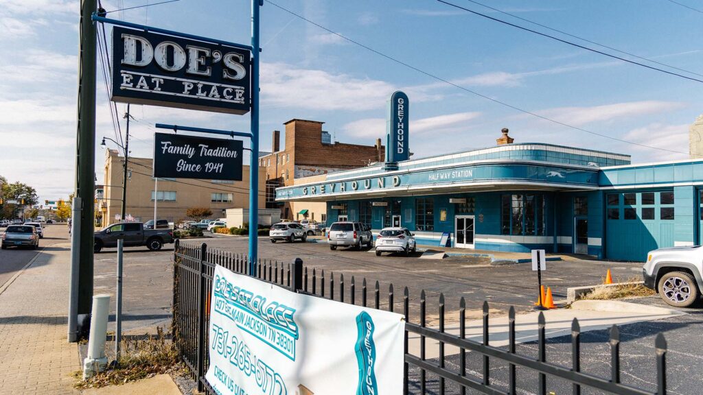 A street view of Doe’s Eat Place sign near a turquoise building labeled “Greyhound Bus Station.” Cars are parked nearby, and there is a banner and black fence in the foreground. The sky is mostly clear.