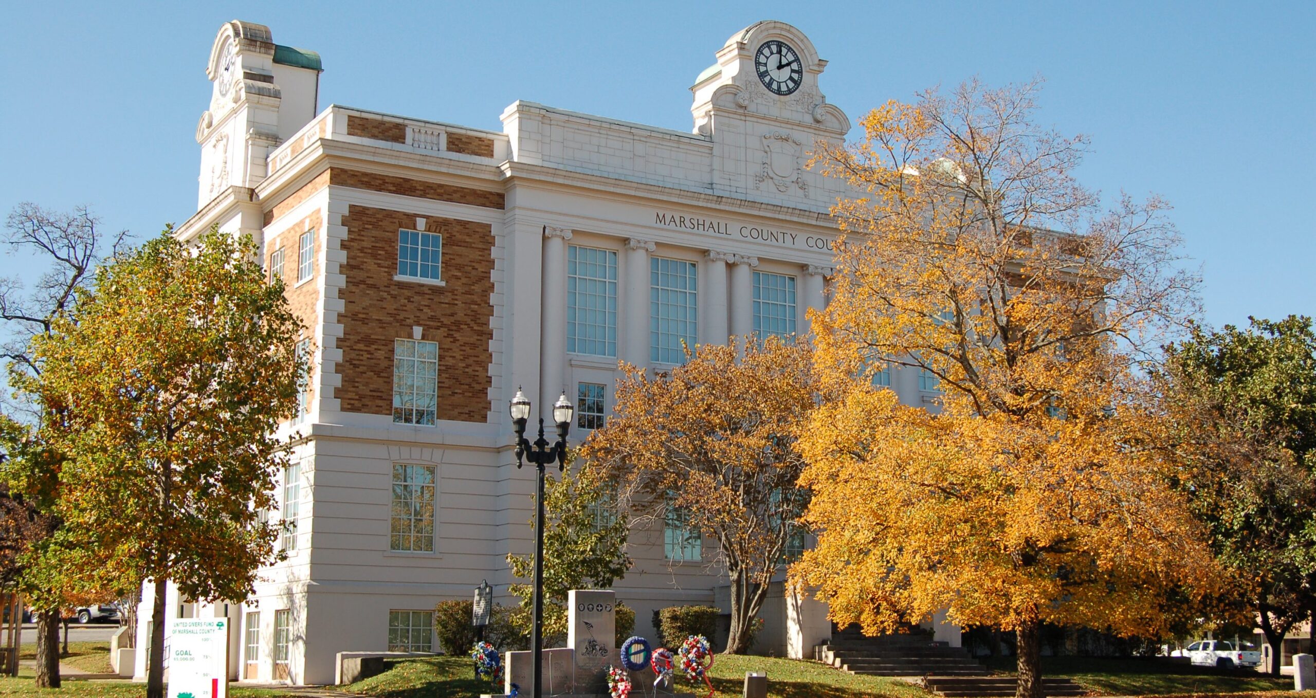 A historic courthouse building with a clock tower and large windows stands behind autumn trees with yellow and green leaves. The name "Marshall County Courthouse" is visible above the entrance.