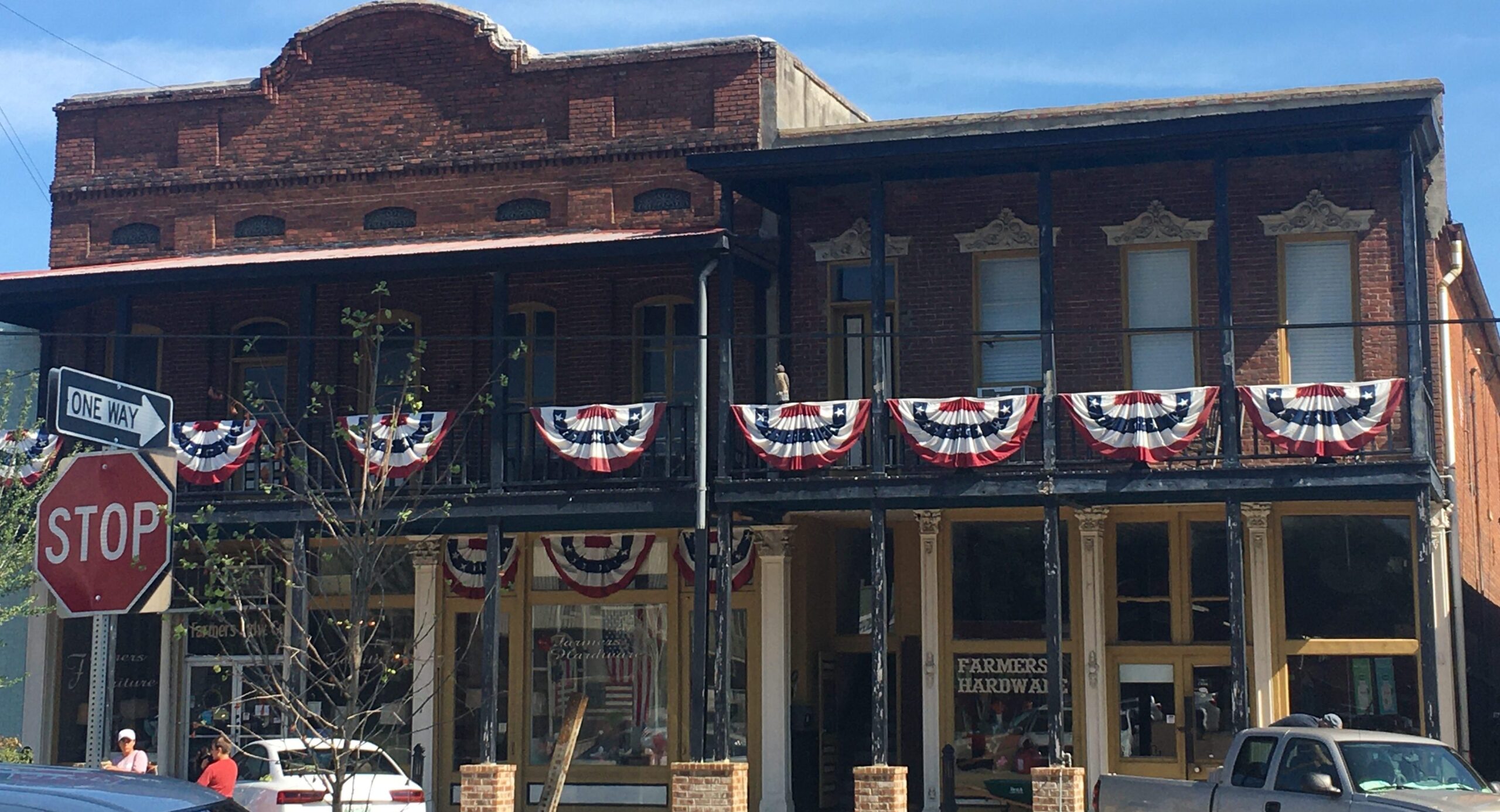 A historic brick building decorated with red, white, and blue bunting. There is a stop sign and one-way street sign in front, and an old-fashioned Farmers Hardware store on the ground floor.