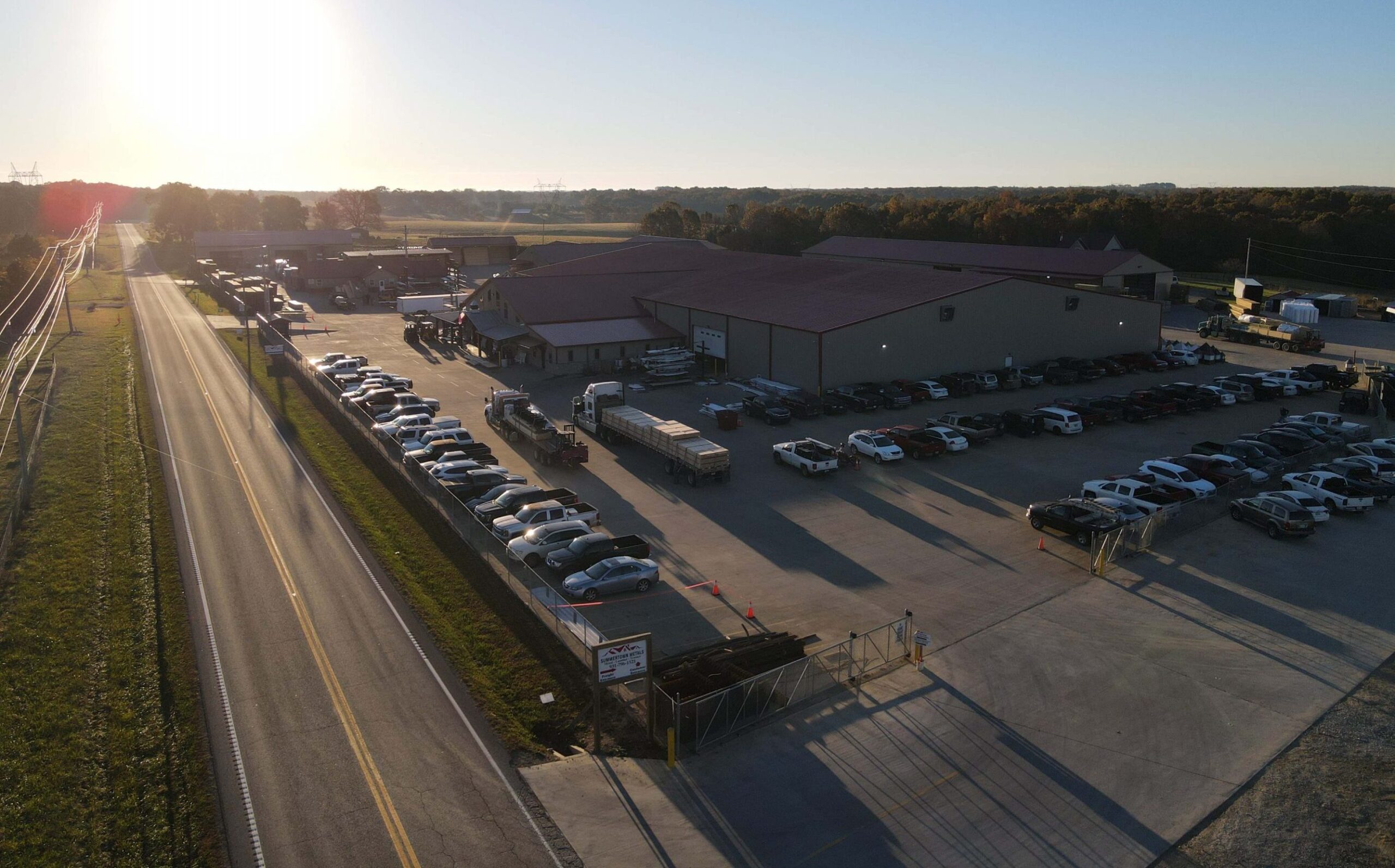 Aerial view of a large industrial building with a parking lot full of cars and trucks, situated next to a road at sunrise or sunset, with fields and trees in the background.