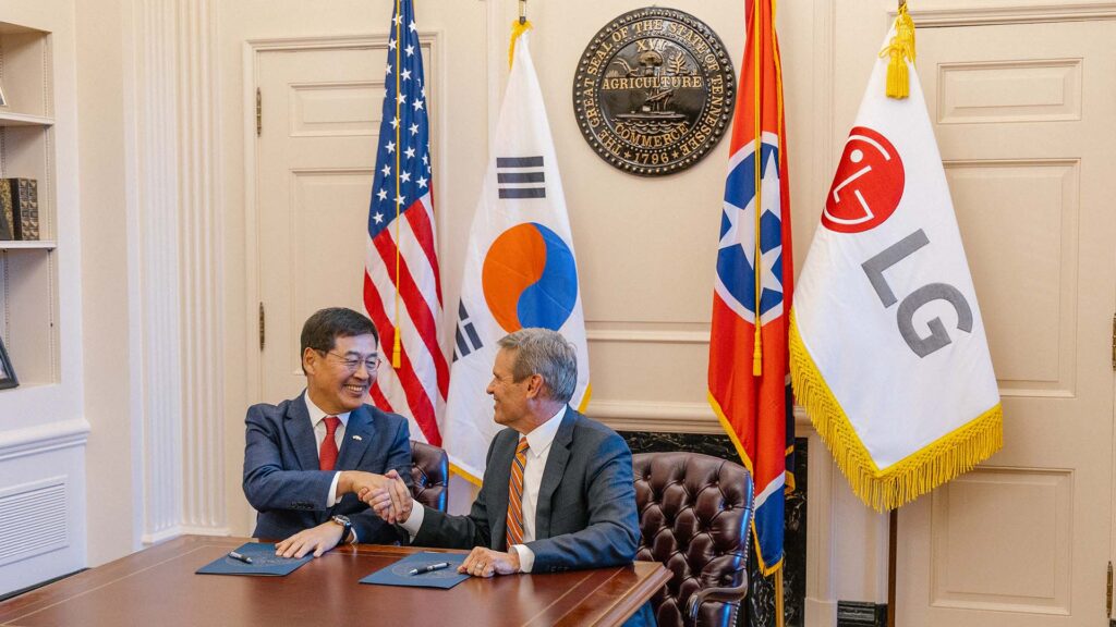 Two men in suits shake hands across a desk with documents, in front of the US, South Korean, Tennessee, and LG flags, inside an official-looking office. A large seal hangs on the wall behind them.