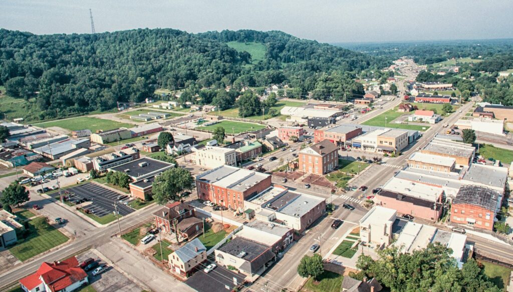 Aerial view of a small town with tree-covered hills in the background, intersecting streets, brick buildings, houses, parking lots, and greenery throughout the area.