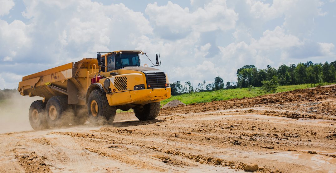 A large yellow dump truck drives on a muddy construction site, kicking up dust under a partly cloudy sky with green trees and grass in the background.