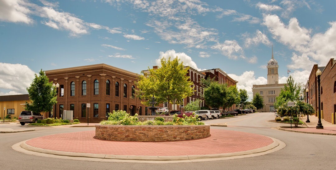 A small roundabout with a raised brick planter and green trees is surrounded by historic brick buildings and parked cars under a partly cloudy blue sky in a quiet downtown area.