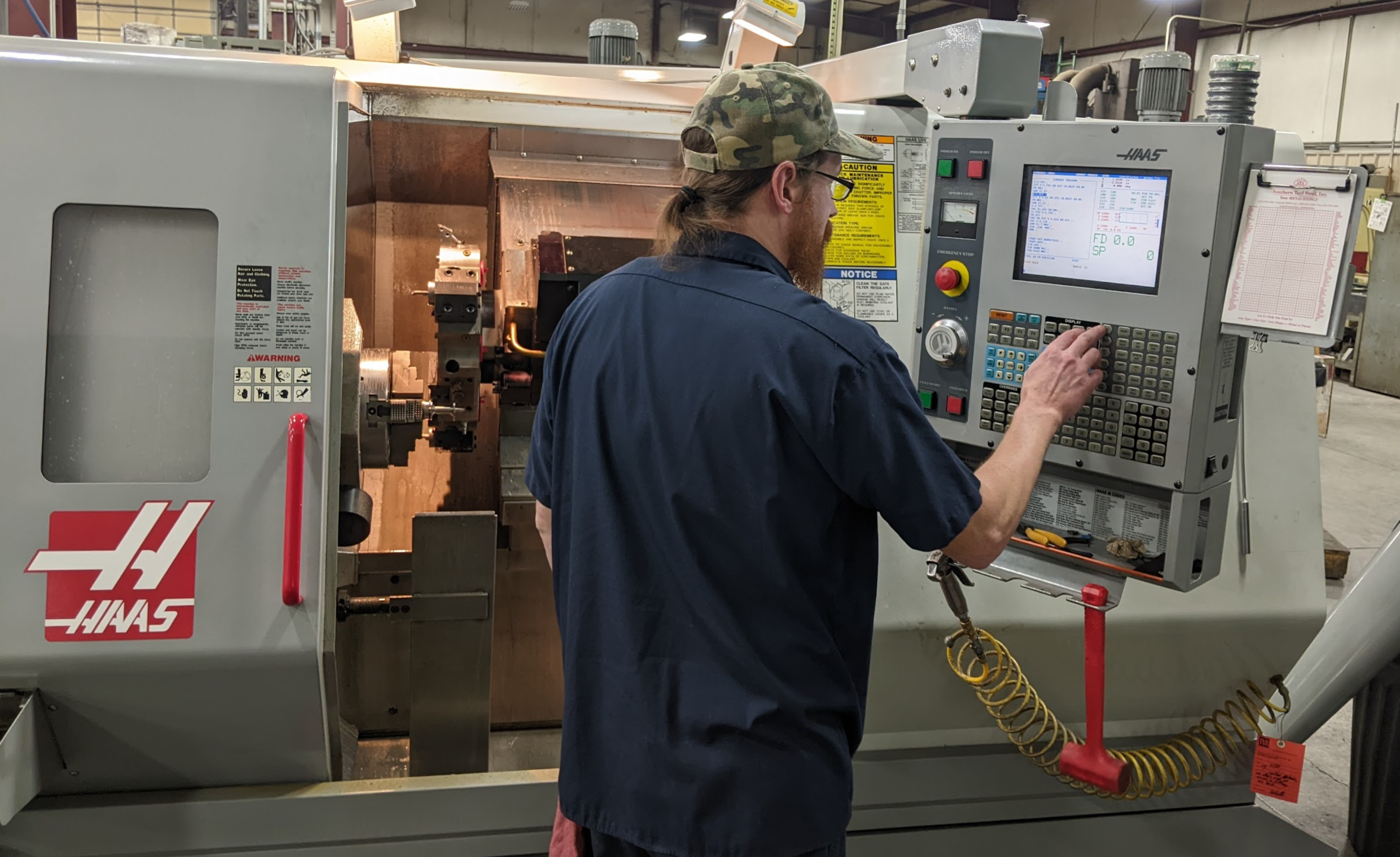 A person in a navy shirt and camouflage cap operates a Haas CNC machine in a workshop, pressing buttons on the control panel while monitoring the screen.
