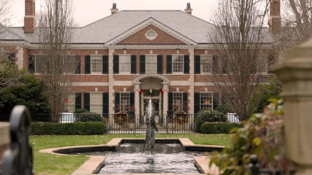 A large brick mansion with black shutters and white trim, featuring a central arched entrance. In front, a fountain sits in a long rectangular pool, surrounded by manicured shrubs and trees.