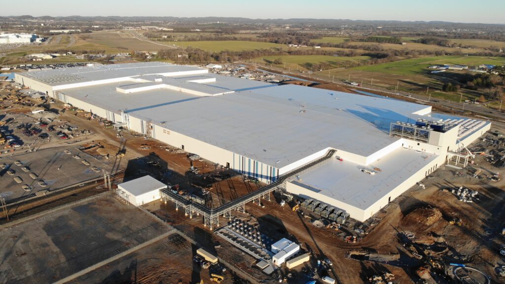 Aerial view of a large industrial building under construction, surrounded by dirt, construction materials, vehicles, and open fields on a clear day.