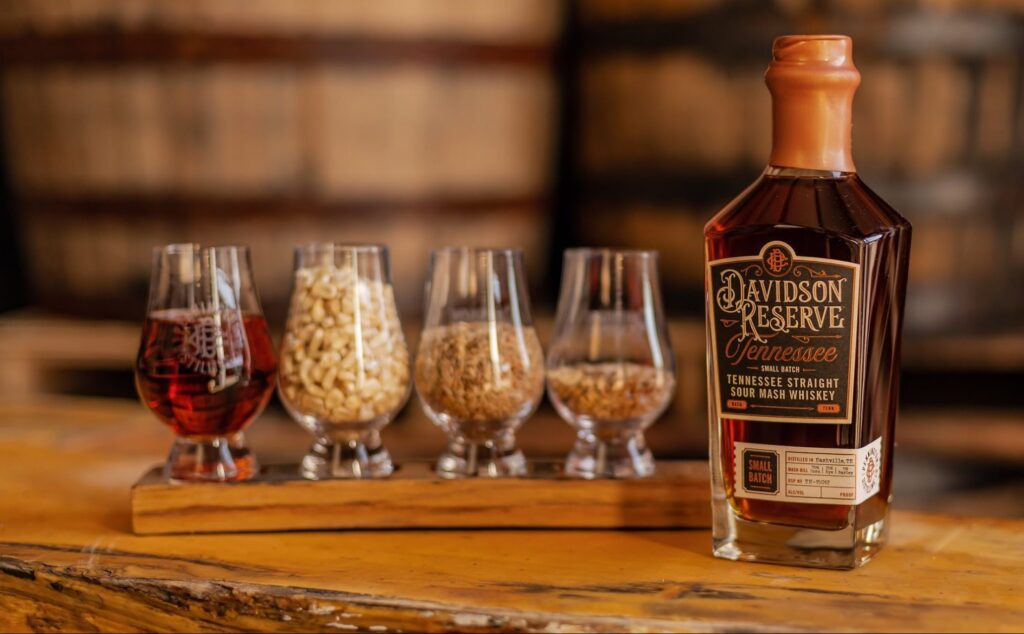 A bottle of Davidson Reserve Tennessee Straight Sour Mash Whiskey stands next to four glasses containing red liquid and various grains, all set on a wooden surface with large barrels in the blurred background.