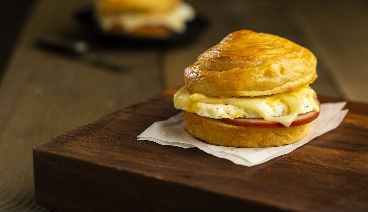A breakfast sandwich with a biscuit bun, egg, melted cheese, and a slice of tomato sits on parchment paper on a wooden surface. Another sandwich is blurred in the background.