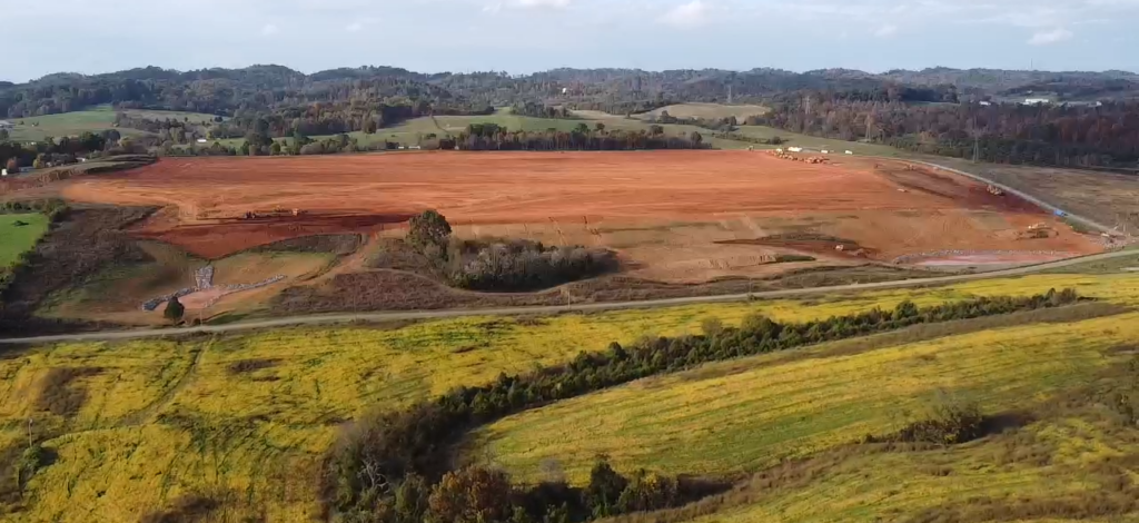 A wide view of rolling hills with patches of yellow-green grass in the foreground and a large, flat area of exposed red soil in the middle, surrounded by trees and distant hills under a partly cloudy sky.
