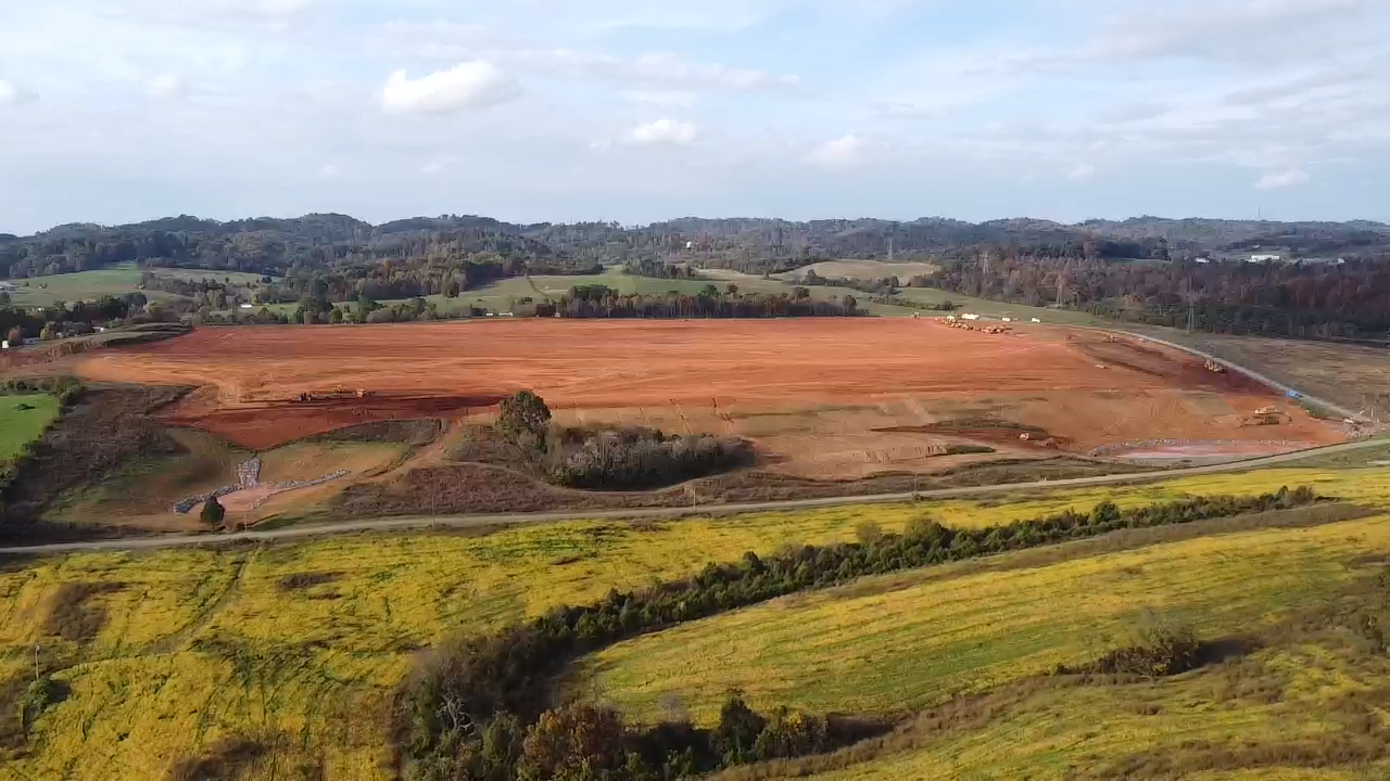A wide view of a rural landscape shows a large, cleared area of red soil in the center, surrounded by green fields and distant hills under a partly cloudy sky.