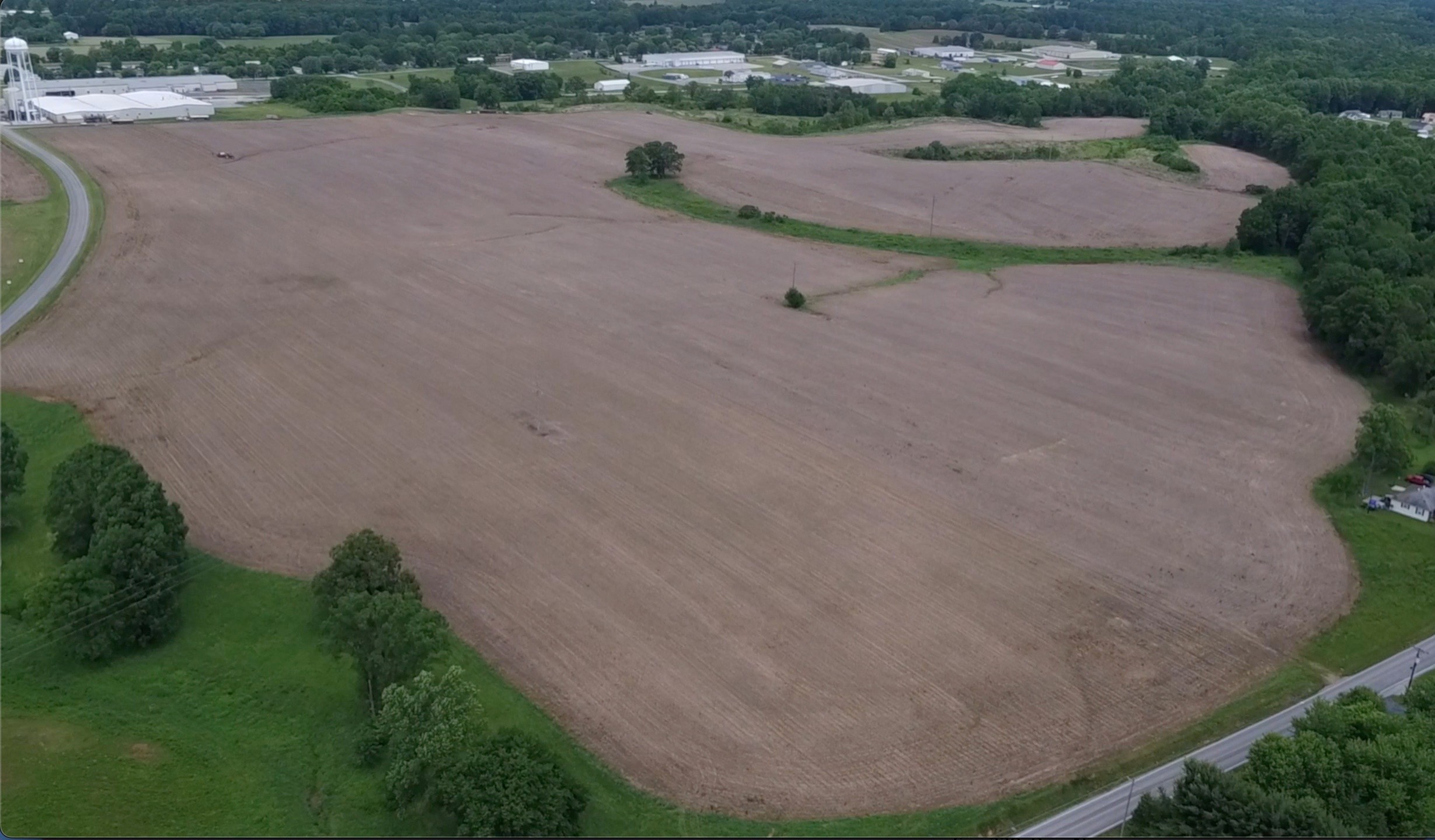 Aerial view of a large, plowed, brown field bordered by green trees and grass, with some buildings and forest visible in the background under a cloudy sky.