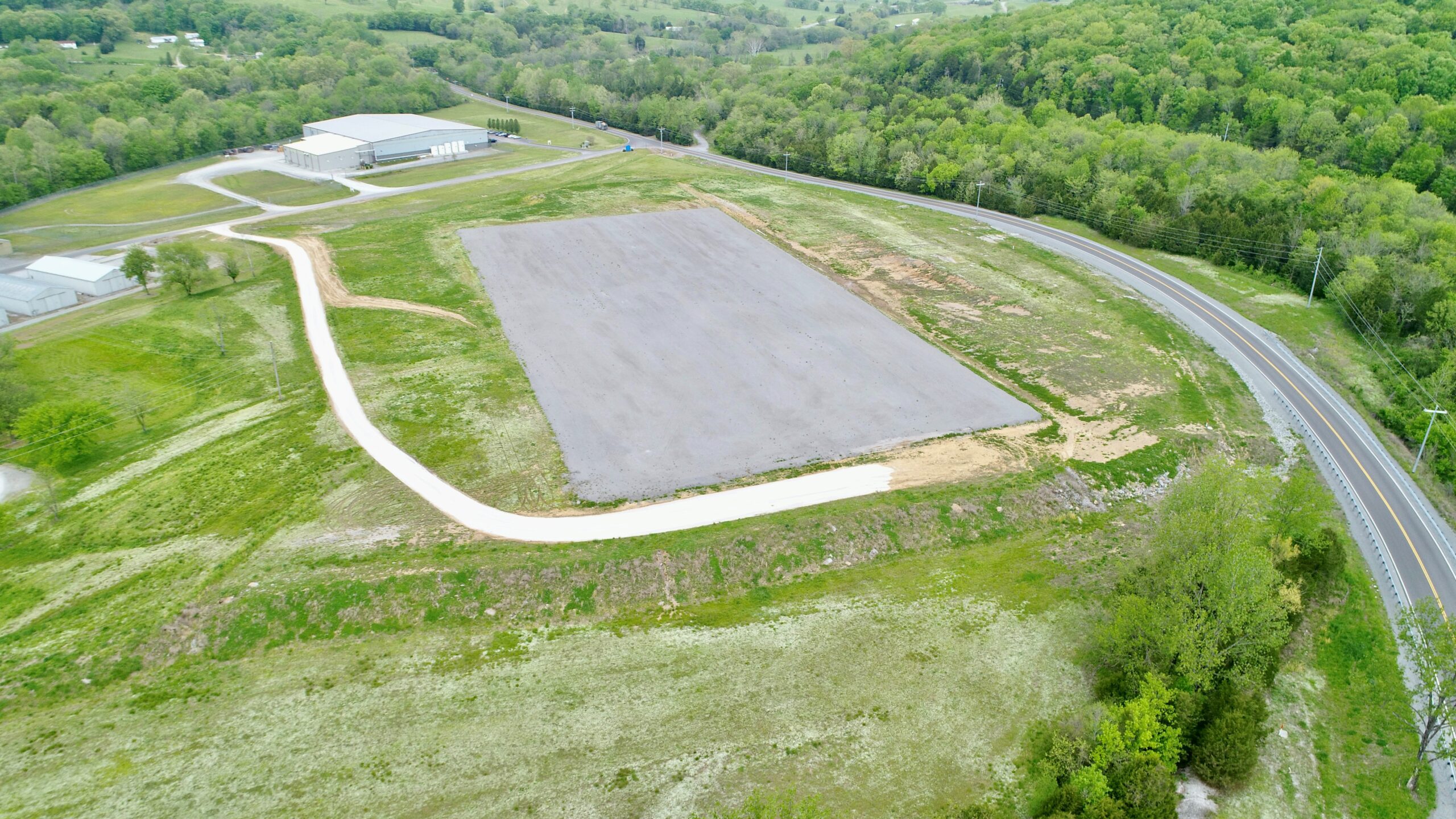 Aerial view of a large rectangular paved lot surrounded by grassy fields and trees, with a curving dirt road leading to it. A building and winding road are visible in the upper left and right areas of the image.