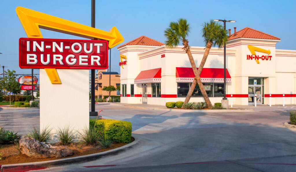 A drive-thru In-N-Out Burger restaurant with palm trees outside, red and yellow signage, and a clear blue sky in the background.