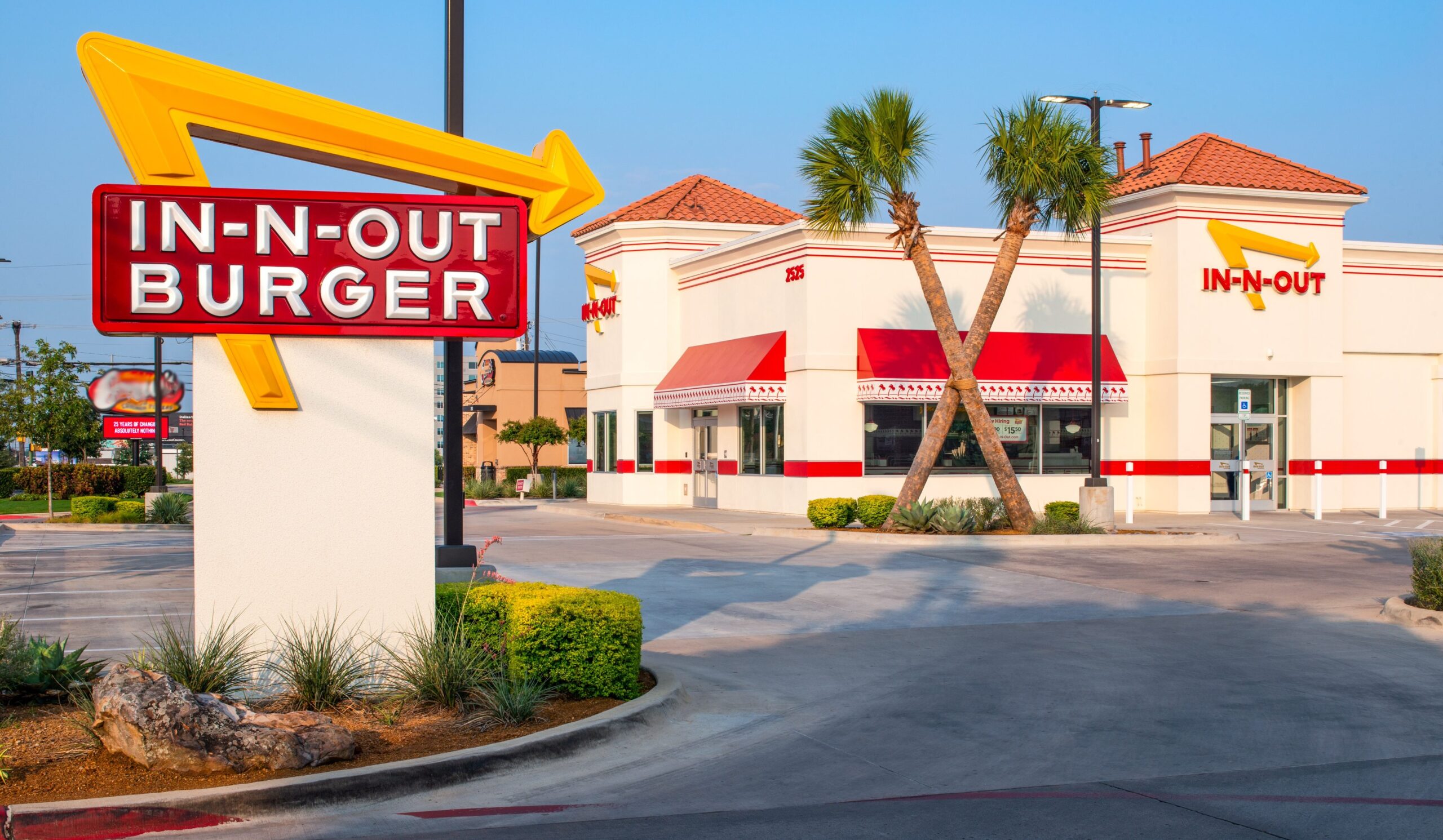 A drive-thru In-N-Out Burger restaurant with palm trees outside, red and yellow signage, and a clear blue sky in the background.