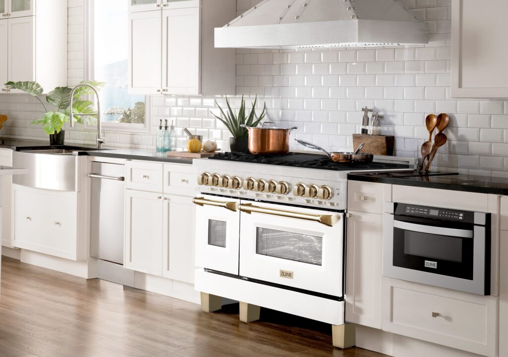 A modern kitchen with white cabinets, stainless steel appliances, a white tile backsplash, a farmhouse sink, potted plants, and a large white and gold range with a double oven beneath a silver vent hood.